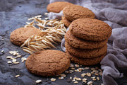 Stack of brown ragi biscuits on a dark surface with scattered oats and rice straws, partially covered by a grey napkin.